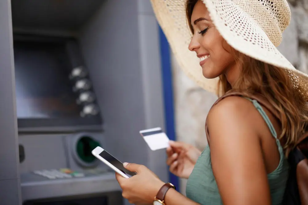 An OnlyFans creator smiling while using a smartphone and debit card to withdraw cash from an ATM.