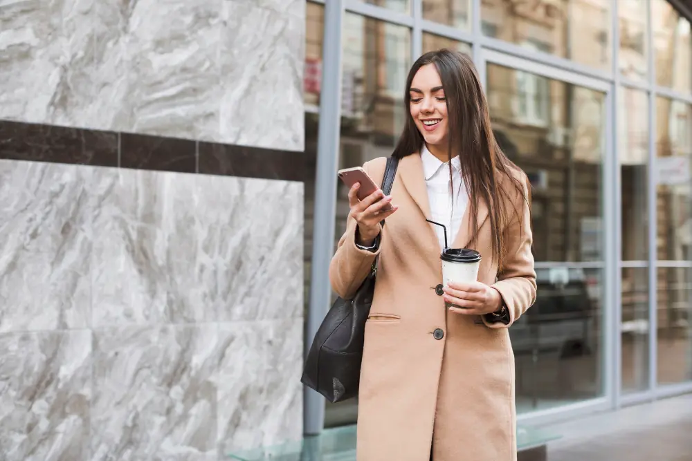 Woman in a beige trench coat discreetly checking her phone during her morning work commute.